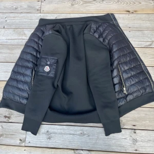 Svart cardigan från Moncler S - Svart pufferjacka från Moncler i storlek S. Jackan har quiltade partier på ärmar och sidor, dragkedjor på fickorna och en snygg Moncler-logga på ärmen. Materialmix med glansig polyester och mjuk bomullskänsla. Perfekt för dig som vill ha en stilren och sportig look.