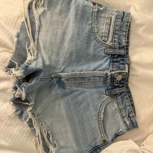 Ljusblå jeansshorts med fransar från Zara - Säljer ett par ljusblå jeansshorts från Zara med snygga slitningar och fransade kanter. Klassisk femficksmodell med bälteshällor och knappstängning. Perfekta för en chill sommarlook.