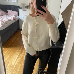 Vit kofta med rosettdetaljer - Supersöt vit kofta med rund hals och knappar framtill. Koftan har unika rosettdetaljer längs knappraden och är stickad i ett mjukt material. Långärmad och ribbade muddar vid ärmslut och nederkant.