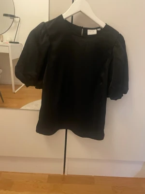 Svart blus med puffärm från Vila - Säljer en svart blus från Vila i storlek 34. Blusen har puffiga korta ärmar och rund halsringning. Materialet är mjukt och har en lätt glansig finish. Perfekt för dig som gillar stilrena och enkla plagg med en twist. Jag säljer denna topp/blus, då den tyvärr är för liten och kommer inte till användning längre.