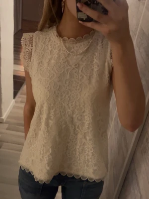 Vit spetsblus med scallopkant - Säljer en söt vit blus i spets med scallopkant vid hals och nederkant. Blusen är ärmlös med fina spetsdetaljer på axlarna och har ett romantiskt mönster över hela plagget. Perfekt att styla med jeans eller kjol för en fräsch look.