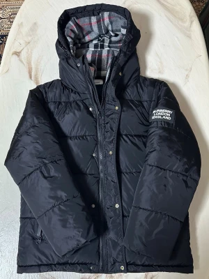 Svart pufferjacka från Burberry - Svart pufferjacka från Burberry med huva och dragkedja framtill. Insidan har ett rutigt foder i grått, vitt, svart och rött. Jackan har en patch med Burberry London England på ärmen och praktiska fickor med dragkedja. Perfekt för kalla dagar och riktigt snygg stil.