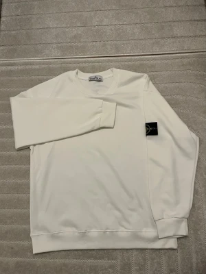 Vit sweatshirt från Stone Island - Vit långärmad sweatshirt från Stone Island i mjuk bomull. Klassisk rund hals och ribbade muddar vid ärmslut och nederkant. Ikonisk svart patch med gul och grön broderad logga på vänster ärm. Perfekt för en clean och stilren look. Varan har inte använts alls. 