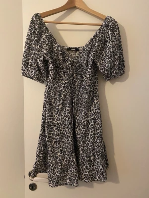 Blommig kortklänning från Bik Bok - Supersöt kortklänning från Bik Bok med svart blommigt mönster på vit botten. Klänningen har puffiga kortärmar, v-ringad hals och snörning framtill. Perfekt för varma dagar och har en lätt och luftig känsla tack vare bomullsmaterialet.