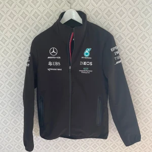 Svart Mercedes Petronas vindjacka - Det Svart vindjacka från Mercedes AMG Petronas med broderade loggor och sponsors på bröst och ärmar. Hög krage med röd/vit/blå detalj, dragkedja framtill, svårt att sätta pris på den här eftersom den är så svår att få tag i och den enda på marknaden, skriv om du skulle va intresserad och kom med prisförslag 