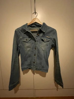 Blå jeansjacka från Tessie - Säljer en klassisk blå jeansjacka från Tessie med snygga bronsfärgade knappar och två bröstfickor med lock. Jackan har en figurnära passform och långa ärmar. Perfekt att slänga över en hoodie eller t-shirt för en chill look.