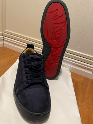 Mörkblå sneakers från Christian Louboutin - Säljer ett par mörkblå sneakers från Christian Louboutin med klassisk röd sula och rund tå. Skorna har snörning och är tillverkade i mocka med svart yttersula. Insidan är beige och mjuk för extra komfort. Perfekt för dig som gillar lyxiga detaljer och stilren design. Box ingår. Priset är inte hugget i sten