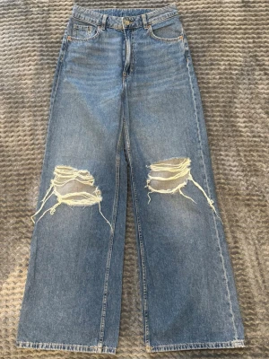 Blå wide jeans med slitna knän H&M Divided - Säljer ett par blå wide fit jeans från H&M Divided i storlek 38. Jeansen har hög midja, raka och vida ben samt stora slitningar över båda knäna för en riktigt edgy look. Klassisk femficksmodell i 100% bomull.