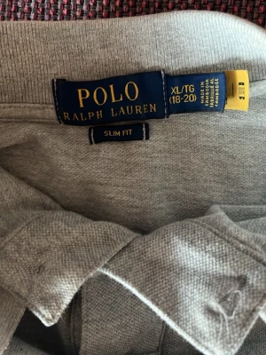 Grå polotopp från Polo Ralph Lauren - Säljer en grå polotopp från Polo Ralph Lauren i slim fit-modell. Toppen har korta ärmar, klassisk krage och två knappar framtill. Materialet är mjukt och bekvämt, perfekt för en chill och stilren look. Diskret logga broderad på bröstet.