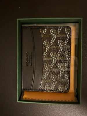 Svart G plånbok med mönster - Säljer en stilren svart plånbok från G med ikoniskt grafiskt mönster i vitt och grått. Plånboken är tillverkad i skinn och har flera fack för kort. Kommer i original grön ask med logga. Perfekt för dig som gillar lyxiga accessoarer.