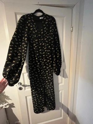 Svart blommig långklänning från Adoore - Säljer en svart långklänning från Adoore med små blommiga mönster i ljusgult. Klänningen har v-ringad hals, långa puffärmar och knappar framtill. Perfekt för t ex nyår eller andra festligheter! ✨ Skulle du önska att ha den nu till nyår kommer jag absolut prioritera att posta samma dag! 