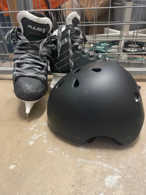 Svarta skridskor Pulse 5 med hjälm - Säljer ett par svarta skridskor från Pulse 5 med vit och grå fodring, samt svart och vitmönstrade snören. Skridskorna har rund tå och platt sula. Medföljer en svart hjälm med ventilationshål och justerbart spänne. Perfekt för vinterns isaktiviteter. Både skridskorna och hjälmen för endast 70kr. 