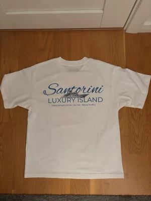 Vit Maison De Rêve t-shirt Santorini - Säljer en fräsch vit t-shirt från Maison De Rêve med tryck på både fram- och baksidan. På ryggen står det 'Santorini Luxury Island' i blått och framtill finns en mindre logga. T-shirten har rund hals och korta ärmar, perfekt för chill dagar. Aldrig använd. Lite oversized