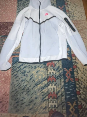 Vit vindjacka från Nike med huva - Snygg vit vindjacka från Nike med svart dragkedja och huva. Jackan har en cool detalj med svart linje över bröstet och en rosa Nike-logga. Långärmad och med ficka på ärmen, perfekt för en sportig stil.