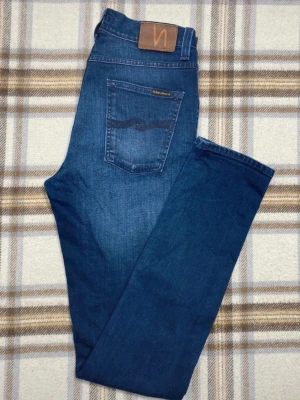 Mörkblå jeans från Nudie Jeans Co - Snygga mörkblå jeans från Nudie Jeans Co i klassisk straight fit. Jeansen har fem fickor, subtila slitningar och Nudie-logga på bakfickan. Materialet är slitstarkt jeans och passformen är normal. Perfekt för dig som gillar stilrena och tidlösa jeans.