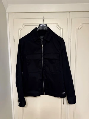 Svart overshirt från Zara med dragkedja - Svart overshirt från Zara med klassisk krage och två stora bröstfickor. Jackan har en snygg silverfärgad dragkedja framtill och är tillverkad i ett slitstarkt bomullsmaterial. Perfekt för lager-på-lager och enkel att styla till olika outfits.