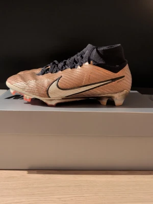 Nike Mercurial Vapor 16 Elite fotbollsskor - Säljer ett par Nike Mercurial Vapor 16 Elite KM AG-Pro i metallic kopparfärg med svarta detaljer och vit swoosh. Skorna har snörning, hög stickad strumpkrage och platt sula med dobbar för konstgräs. Perfekta för dig som vill sticka ut på planen.