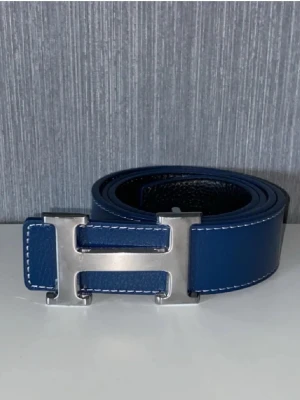 Blå bälte  - Bilderna visar ett blått skärp med en stor H-formad metallspänne, inte en väst. Skärpet har vita sömmar och insidan är svart.