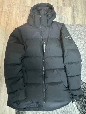 Svart Sail Racing pufferjacka - Säljer en svart pufferjacka från Sail Racing med huva och snyggt Sail Antarctica Expedition-märke på ärmen. Jackan har lång ärm, dragkedja framtill och är perfekt för kalla dagar. Den har en clean look och är riktigt skön att bära. Ny pris 3500kr