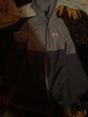 Grå och svart vindjacka från Under Armour - Snygg tvåfärgad vindjacka från Under Armour i grått och svart. Jackan har huva med snörning, lång ärm och dragkedja framtill. Perfekt för dig som gillar sportig stil och vill ha något som står emot vind och väder. Använd 2 gånger bara. Kan gå ner i pris vid snabb affär. Dragkedjan kan krångla lite!