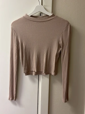Beige ribbad croppad långärmad topp - Säljer en beige långärmad topp med ribbad struktur och croppad passform. Toppen har en enkel, rundad hals och är tillverkad i mjukt material som sitter skönt mot kroppen. Perfekt att styla med jeans eller kjol för en chill look.