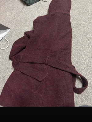 Vinröd ullkappa med huva från Amisu - Säljer en vinröd kappa från Amisu med huva, bälte i midjan och dubbla knapprader framtill. Kappan är tillverkad i ullblandning och har ett svart, slätt innerfoder. Perfekt för dig som gillar en chill och snygg look under höst och vinter.