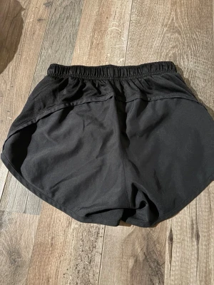 Svarta Nike träningsshorts XS - Nike träningsshorts i svart med elastisk midja och två silvriga swoosh-loggor framtill. Modellen är loose fit och har rundade kanter för extra rörelsefrihet. Materialet är lätt och syntetiskt, perfekt för sport och aktivitet.
