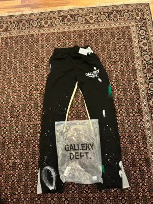 Svarta Gallery Dept. mjukisbyxor - Svarta mjukisbyxor från Gallery Dept. med vit logotyp framtill och konstnärliga färgstänk i vitt och grönt. Byxorna har elastisk midja och snörning, samt kontrastpaneler i beige och grått längs benen. Materialet är mjukt och bekvämt, perfekt för chill dagar. Helt nya