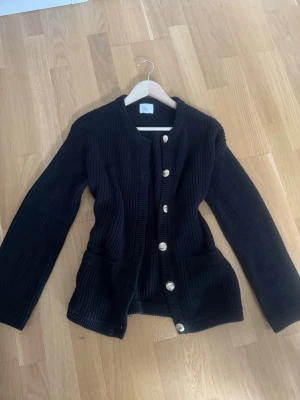 Svart stickad kofta från H&M XS - Säljer en svart, ribbstickad kofta från H&M i storlek XS. Koftan har rund hals, långa ärmar och snygga silvriga knappar framtill. Två fickor framtill ger extra stilpoäng. Perfekt för lager-på-lager och chill dagar.