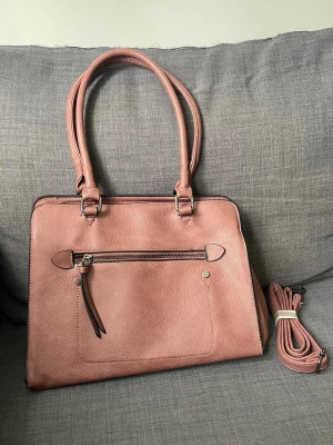 Rosa handväska med axelrem - Säljer en snygg rosa handväska i skinnimitation med tillhörande axelrem. Väskan har praktiska handtag, flera fack med dragkedja och metall-detaljer. Perfekt storlek för att få plats med allt du behöver och passar till många olika stilar.
