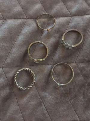 Fem silvriga ringar med olika design - Säljer ett set med fem silvriga ringar i olika stilar. En ring har tvinnad design, en med hjärtformad öppning, två med små stenar och en enkel slät ring. Perfekt för att mixa och matcha till din egen stil. Alla ringar är i metall med silverfärg. Använda fåtal gånger🥰man får alla för 60 kronor 