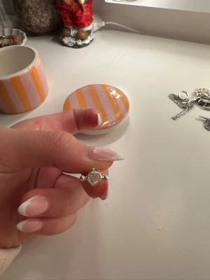 Söta silvriga örhängen med hjärta - Ett par små örhängen i silverfärgad metall med ett hjärtformat motiv i mitten. Hjärtdetaljen har en glittrande sten som ger extra shine. Perfekta om du gillar smycken med gulliga detaljer och vill ha något som sticker ut lite.