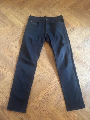 Svarta Diesel jeans, storlek 32 - Säljer ett par svarta jeans från Diesel i storlek 32. Modellen har klassisk femficksdesign, taperd fit och diskreta detaljer med röd Diesel-logga på bakfickan. Materialet är 98% bomull och 2% elastan för skön stretch. Tillverkade i Italien.