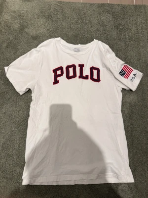 Vit t-shirt från Polo Ralph Lauren - Säljer en asfet T-shirt från Polo Ralph Lauren i bra skick. Den har ett litet hål nere på framsidan och några små fläckar men syns inte vid användning, men därav priset. Den har en amerikansk flagga på ärmen och logga på framsidan. Storlek L/14-16 barn, passar XS. 