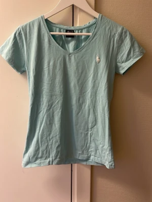 Ljusblå t-shirt från Polo Ralph Lauren - Snygg ljusblå t-shirt från Polo Ralph Lauren med klassisk v-ringning och korta ärmar. T-shirten har en broderad logga på bröstet och är gjord i mjuk bomull som känns skön mot huden. Perfekt för dig som gillar stilrena och enkla plagg.