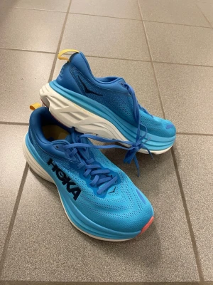 Hoka Bondi 8 blå löparskor EU 40 - Säljer ett par Hoka Bondi 8 sneakers i klarblå färg med vit och rosa sula. Skorna har mesh-ovandel, snörning och Hoka-logga på sidan. Mellansulan är extra tjock för maxad dämpning och komfort. Perfekta för dig som gillar att springa långt och vill ha riktigt sköna skor.