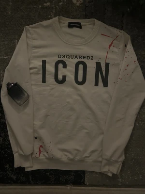 Vit sweatshirt från Dsquared2 ICON - Säljer en vit sweatshirt från Dsquared2 i storlek S med stor svart ICON-print på bröstet och röda samt svarta färgstänk som detaljer. Tröjan har rund hals, ribbade muddar och är långärmad. Perfekt statement-plagg för dig som gillar streetwear.