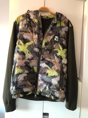 Camouflage pufferjacka från Etro - Säljer en unik pufferjacka från Etro med camouflage-mönster i grönt, beige och inslag av gult. Jackan har stickade, olivgröna ärmar och huva samt dragkedjor framtill och på fickorna. Märkeslogga på bröstet och cool mix av material för en modern look.