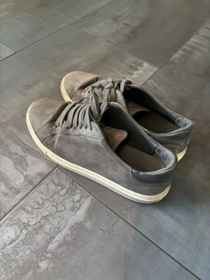 Mocka Arigato skor - Säljer ett par grå sneakers med ovandel i mocka och matchande grå snören. Skorna har en vit platt sula och rund tå. Baktill finns en mörkare detalj i materialet. Perfekt för dig som gillar stilrena och enkla sneakers. Väl använda och säljes därför till lägre pris!