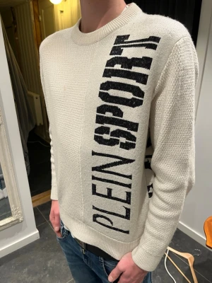Vit stickad tröja från Plein Sport - Säljer en vit stickad tröja från Plein Sport i storlek M. Tröjan har ett grovt stickat mönster, svart grafiskt tryck med texten 'PLEIN SPORT' framtill och en stor svart tigerdesign på ryggen. Rund hals och långa ärmar, perfekt för dig som gillar streetwear.