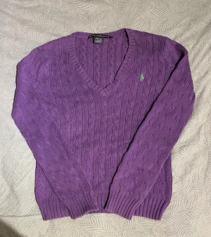 Lila stickad tröja från Ralph Lauren - Säljer en lila kabelstickad tröja från Ralph Lauren med v-ringad hals och långärmad design. Tröjan har ett grönt broderat logotyp på bröstet och ribbade muddar vid ärmslut och nederkant. Perfekt för dig som gillar klassisk stil med färg.