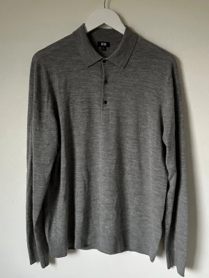 Grå långärmad pikétröja från Uniqlo - Säljer en stilren grå långärmad pikétröja från Uniqlo i storlek L. Tröjan är tillverkad i 100% ull och har klassisk krage med tre knappar framtill. Perfekt för dig som gillar enkel och clean look med mjukt material. 5M5