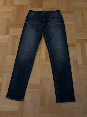 Jack and Jones Jeans 31/34 - Säljer ett par mörkblå jeans från Jack and Jones i modellen Tapered Mike och storlek 31/34. Jeansen har klassisk femficksdesign, snygga slitningar och kontrastsömmar. Passformen är tapered med rakare ben och normal midja. Priset går att diskutera!