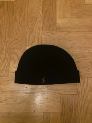 Svart mössa från Polo Ralph Lauren - Svart mössa från Polo Ralph Lauren med klassisk broderad logga i orange framtill. Mössan är ribbstickad och tillverkad i mjuk bomull, perfekt för kalla dagar. Enkel och stilren design som passar till det mesta.