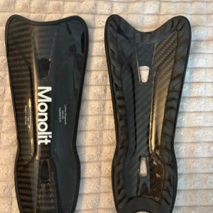 Monolit benskydd i kolfiber - Säljer ett par svarta benskydd från Monolit i kolfiber. De är lätta, tunna och har en snygg glansig finish med vit logga. Kommer i ett smidigt fodral med skuminsats för säker förvaring. Perfekta för dig som vill ha proffsig utrustning på planen.