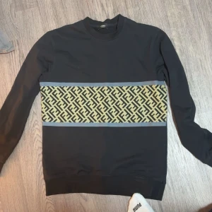 Svart sweatshirt med mönstrad panel - Svart långärmad sweatshirt från Kenzo med en bred panel över bröstet i beige och svart grafiskt mönster. Tröjan har rund hals och ribbade muddar vid ärmslut och nederkant. Perfekt för dig som gillar stilrena detaljer och coola prints.
