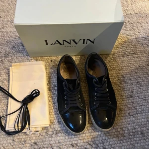 Svarta sneakers från Lanvin med lackdetalj - Snygga svarta sneakers från Lanvin med klassisk rund tå och cool lackad tådel. Skorna har snörning, vit sula och är tillverkade i en mix av mocka och skinn. Perfekta för dig som gillar stilrena men ändå lyxiga sneakers. Skokartong och dustbag medföljer.