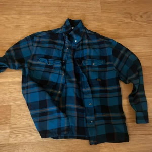 Blå rutig skjorta från H&M - Blå och svart rutig skjorta från H&M i storlek 164. Skjortan har klassisk krage, tryckknappar och två bröstfickor med lock. Perfekt för dig som gillar en avslappnad och cool stil.