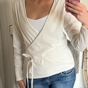Vit omlottopp med knytband - Snygg vit omlottopp med långa ärmar och knytband i midjan. Toppen har en v-ringad halsringning och är tillverkad i ett ribbat material som ger en stilren och fräsch look. Perfekt att matcha med jeans eller kjol för en enkel men trendig outfit.