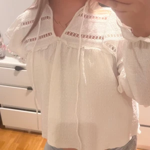 Vit blus med spets och knytband - Säljer en luftig vit blus med långa ärmar, v-ringning och dekorativa spetsdetaljer upptill. Blusen har knytband vid halsen och ett lätt strukturerat tyg som ger en cool vintagekänsla. Perfekt till jeans eller kjol för en fräsch look.💕